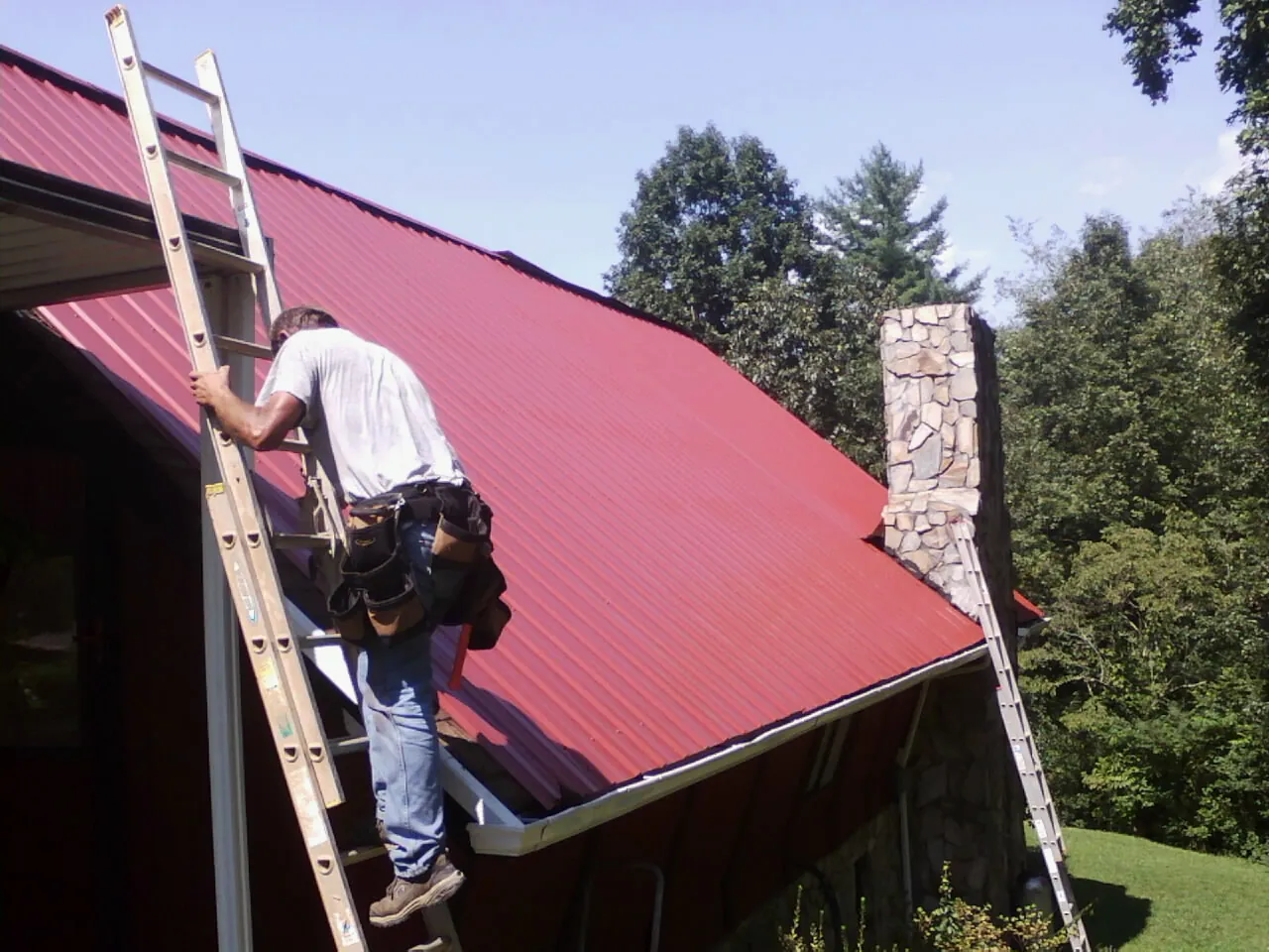 Roofer on a ladder beside a red metal roof and stone chimney — Soffit & Fascia Repair by Roofing Brothers in Westchester